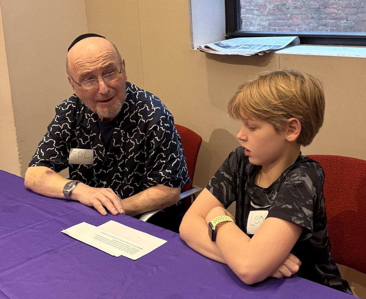 Photo of an older man wearing a yarmulke and a young boy sitting at a table talking.
