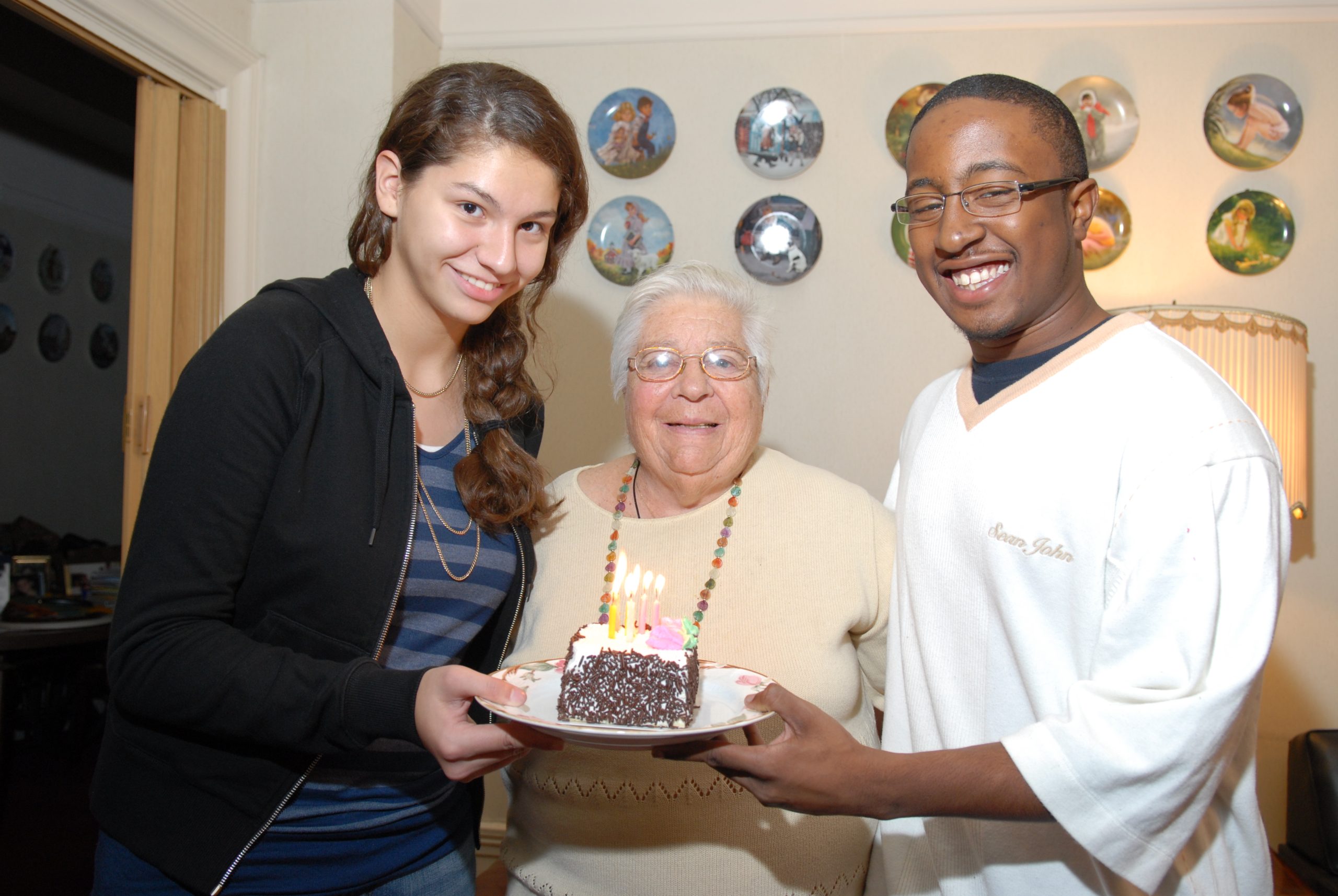Two volunteers holding a piece of birthday cake for an older adult in celebration of her birthday.