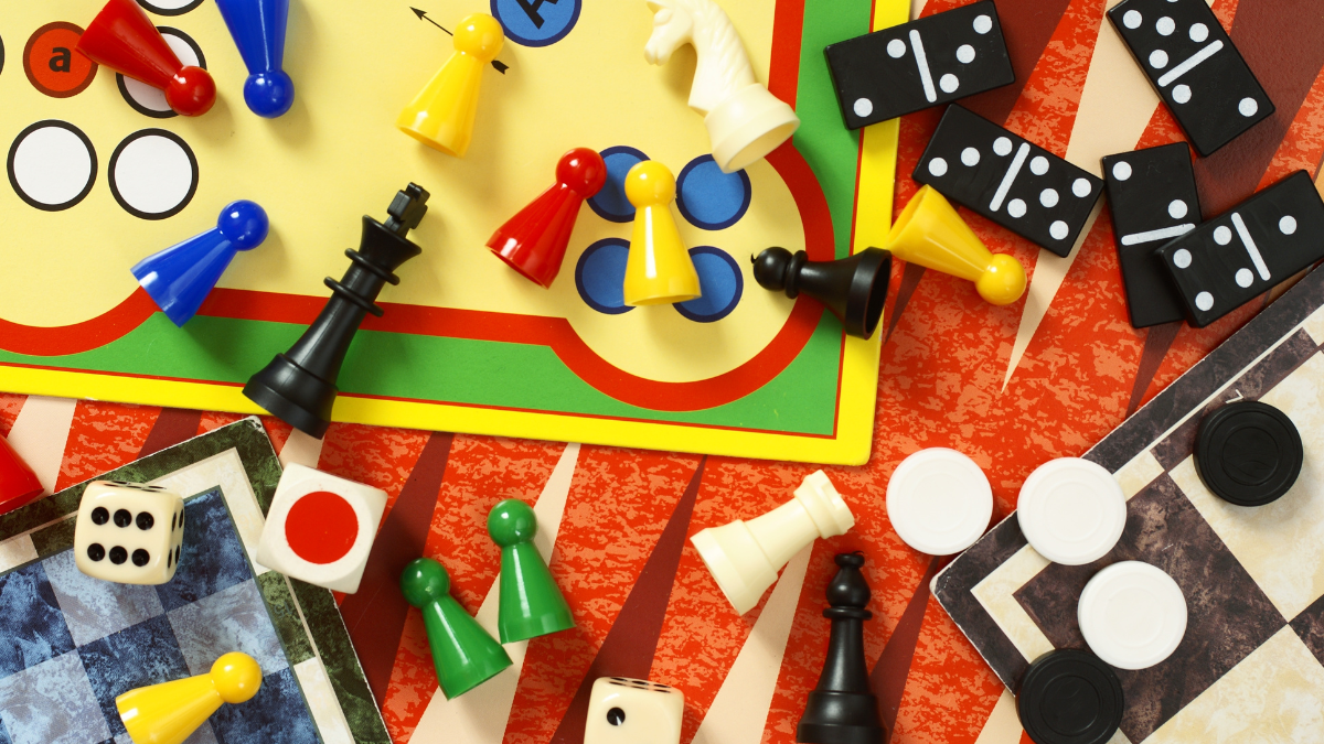 Aerial view of a tabletop covered in board game boards and pieces, including dice, dominoes, checkers, and Sorry game pieces.